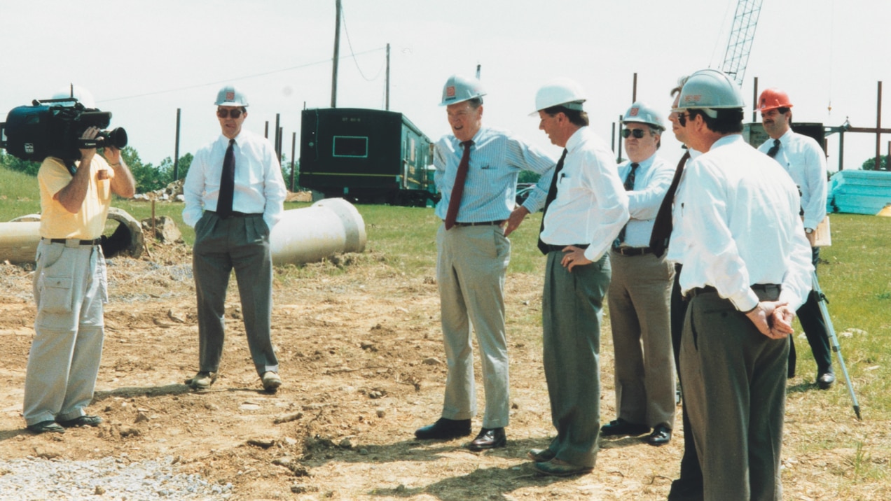 A group of seven men in hardhats stand talking on a construction site. Another man uses a video camera to record them.